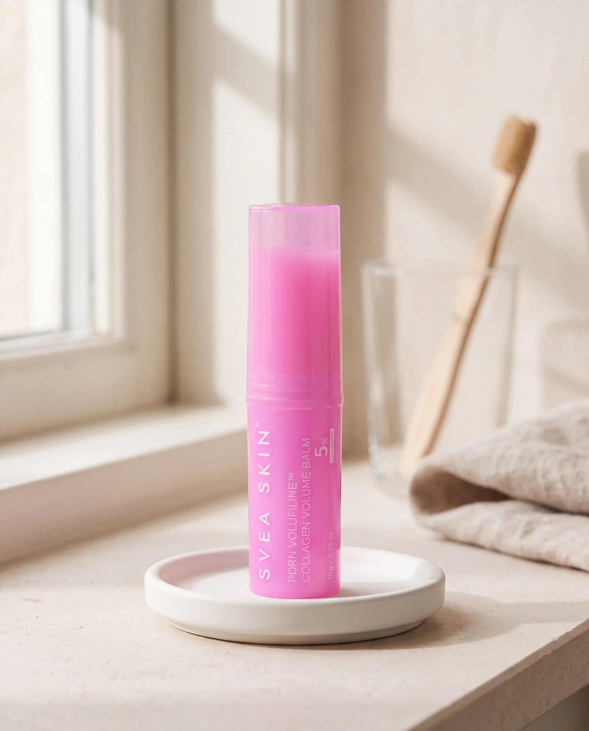 Rosa ansiktsstift från Svea Skin med lock på ett vitt keramikfat på en fönsterbräda i badrummet, bredvid ett glas med en tandborste i trä.