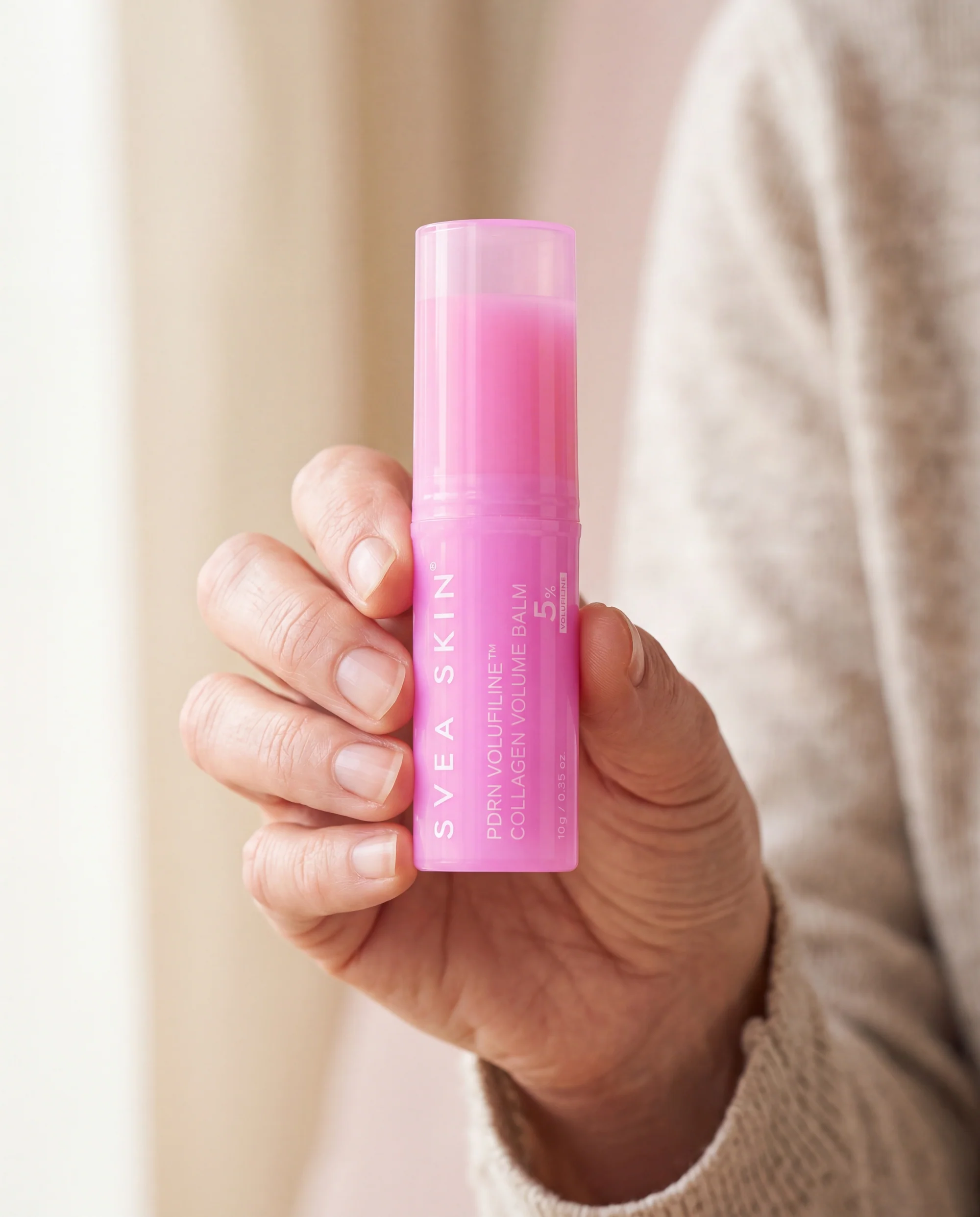 En hand håller upp ett rosa ansiktsstift från Svea Skin framför en beige stickad tröja, så att storleken syns.