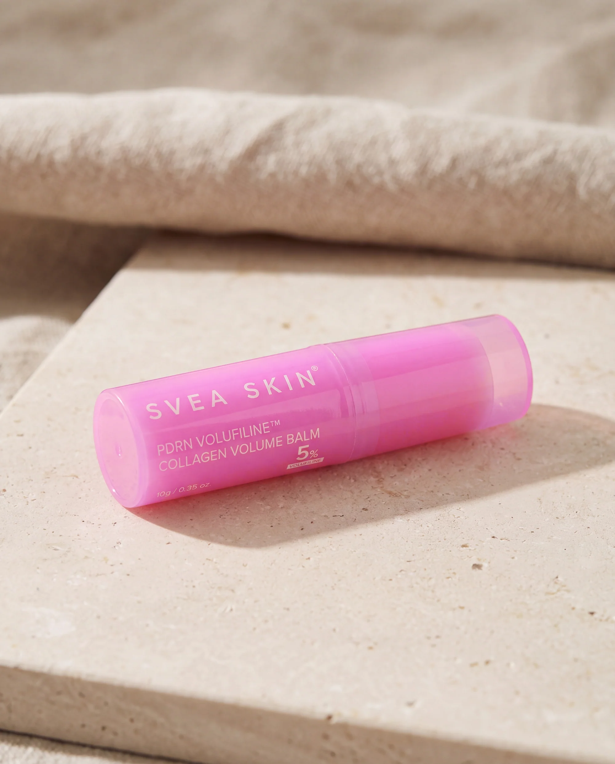 Rosa ansiktsstift från Svea Skin liggande på en ljus kalkstensplatta med ett veckat linnetyg bakom.
